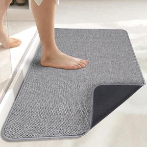 May include: A rectangular bath mat with a textured gray fabric top surface and a solid black rubberized backing. The mat features rounded corners and a stitched gray border, designed to provide a stable surface for stepping out of a shower.