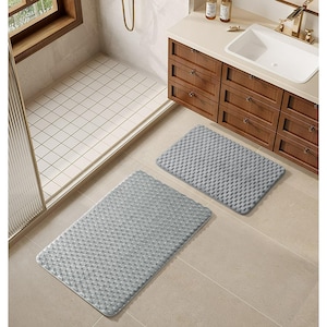 May include: Two gray, rectangular bath mats with a textured, raised pattern. The mats are placed on a tiled floor in a bathroom setting, near a shower and a wooden vanity with a white sink.
