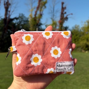 May include: A small, quilted pouch with a daisy pattern in white and yellow on a coral background. The pouch has a zipper closure and a small loop with a key ring. The label reads "Mattison's Stitch Co."