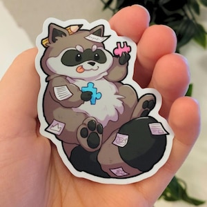 以下が含まれることがあります： グレーと茶色の体、黒いマスク、白と黒の縞模様の尻尾を持つ漫画のアライグマのステッカー。 アライグマはパズルのピースを持っており、小さな紙に囲まれています。 ステッカーには白い縁があります。