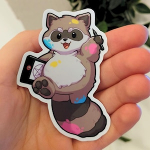 Op de afbeelding: Een sticker met een cartoon wasbeer die een stylus en een tablet met een pentagramontwerp vasthoudt. De wasbeer is bruin, bruin en wit met kleurrijke verfspatten. De sticker staat op een witte achtergrond.