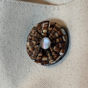 May include: A round button with a mosaic design. The button features a radial pattern of brown and tan shell pieces, with a pearlescent center. The button is set against a neutral fabric background.
