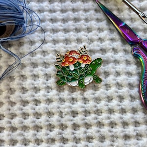 May include: A colorful enamel pin featuring a green turtle with a white belly, adorned with red and white mushroom details and green leaves. The pin has gold-colored outlines. The background includes blue thread and iridescent scissors.