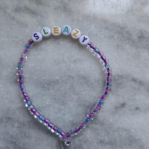 Könnte beinhalten: Ein Perlenarmband mit dem Wort "SLEAZY" in weißen Buchstabenperlen. Das Armband hat eine Mischung aus transparenten, lila und blauen Perlen. Ein kleiner Anhänger hängt am Verschluss.