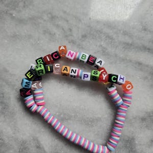 May include: Two beaded bracelets. One bracelet features colorful letter beads spelling "AMERICAN PSYCH". The other bracelet is made of pink, white, and blue disc beads. The bracelets are on a light gray surface.