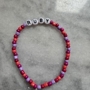 Puede incluir: Una pulsera de cuentas con el nombre "RUBY" escrito con cuentas blancas. La pulsera presenta una mezcla de cuentas rojas, rosas y moradas. Las cuentas están ensartadas para formar una forma circular.