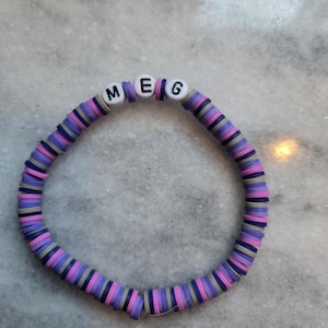 May include: A beaded bracelet with the name "MEG" spelled out in white letter beads. The bracelet features a pattern of purple, pink, and gray disc-shaped beads. The bracelet is circular and rests on a gray surface.