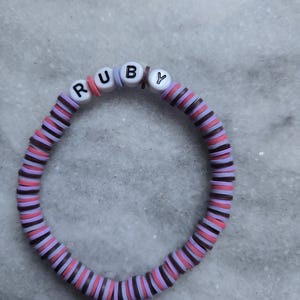 May include: A bracelet made of disc-shaped beads in shades of pink, purple, and brown. The name "RUBY" is spelled out in white letter beads. The bracelet is displayed on a light grey surface.