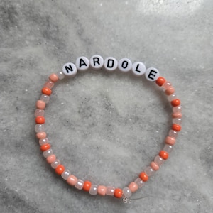 May include: A beaded bracelet with the name "NARDOLE" spelled out in white letter beads. The bracelet features a mix of orange, pink, and clear beads, strung together on a clear elastic cord. The bracelet is displayed on a gray surface.