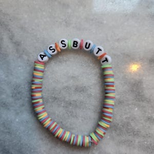 Könnte beinhalten: Ein farbenfrohes Armband mit dem Wort "ASSBUTT" in weißen Buchstabenperlen. Das Armband besteht aus einer Mischung aus pastellfarbenen und hellen Perlen, darunter rosa, blau, grün und orange. Ein lustiges und verspieltes Accessoire.
