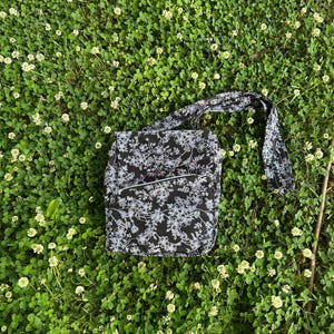 May include: A black and white floral pattern shoulder bag with a matching strap. The bag is rectangular with a front pocket and is lying on a bed of green clover.