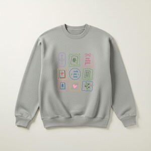 May include: A light gray crewneck sweatshirt featuring a central grid of nine colorful illustrations related to mahjong. The graphic includes mahjong tiles, a pink heart, bamboo stalks, and text that reads "MAHJ", "one more game", and "cracks dots bams".