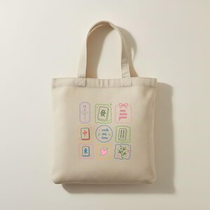 May include: A beige canvas tote bag featuring a colorful grid of nine square and rectangular illustrations. The designs include mahjong tiles, a pink heart, a bamboo stalk, and text that reads "MAHJ", "one more game", and "cracks dots bams".