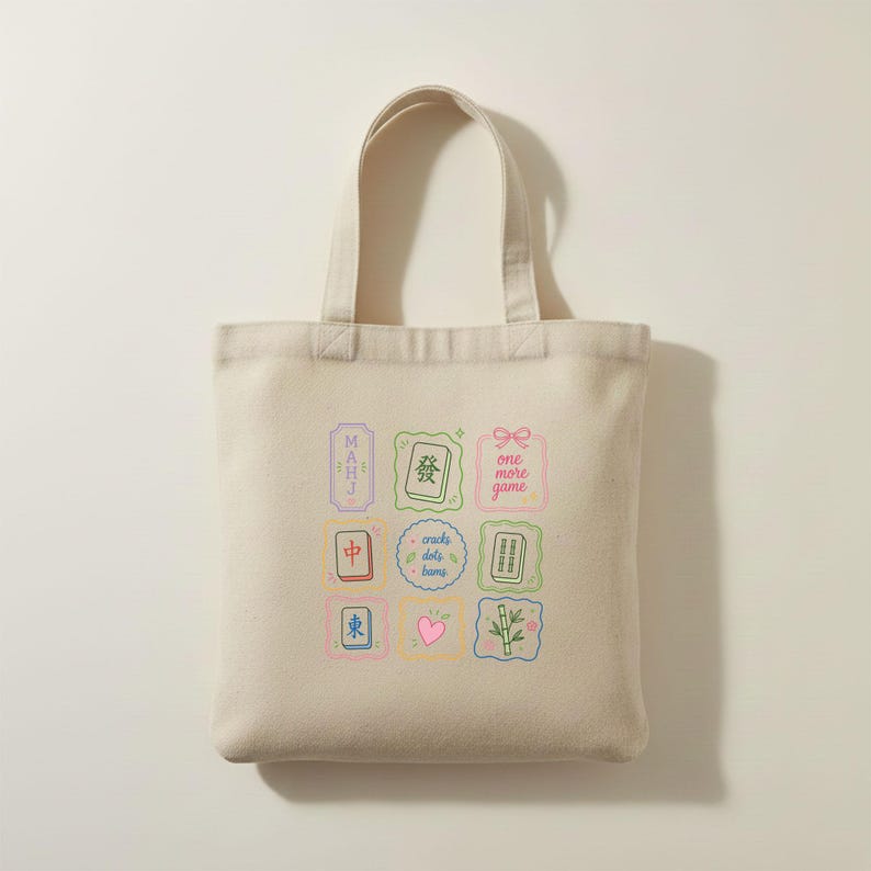 May include: A beige canvas tote bag featuring a colorful grid of nine square and rectangular illustrations. The designs include mahjong tiles, a pink heart, a bamboo stalk, and text that reads "MAHJ", "one more game", and "cracks dots bams".