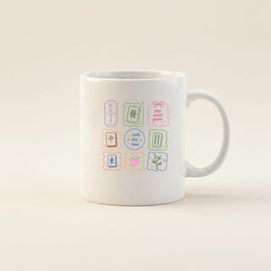 May include: A white ceramic mug featuring a colorful grid of nine square icons related to mahjong. The icons include Chinese characters, bamboo tiles, a pink heart, a bow, and the text "MAHJ" and "one more game" and "cracks dots bams".