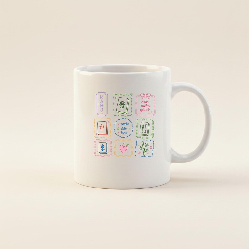 May include: A white ceramic mug featuring a colorful grid of nine square icons related to mahjong. The icons include Chinese characters, bamboo tiles, a pink heart, a bow, and the text "MAHJ" and "one more game" and "cracks dots bams".