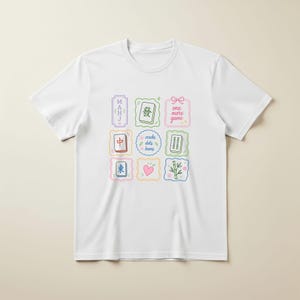 May include: A white short-sleeved t-shirt featuring a colorful graphic print of nine square and rectangular frames containing Mahjong tiles, a pink heart, bamboo stalks, and text that reads "MAHJ", "one more game", and "cracks dots bams".
