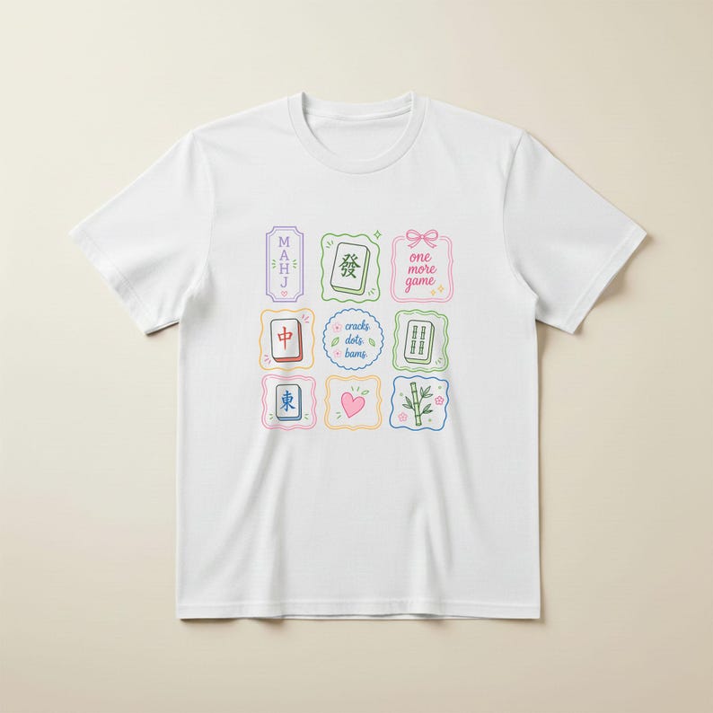 May include: A white short-sleeved t-shirt featuring a colorful graphic print of nine square and rectangular frames containing Mahjong tiles, a pink heart, bamboo stalks, and text that reads "MAHJ", "one more game", and "cracks dots bams".
