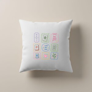 May include: A white square throw pillow featuring a central grid of nine colorful illustrations related to Mahjong. The designs include Mahjong tiles, a pink heart, bamboo stalks, and text that reads MAHJ, one more game, and cracks dots bams.