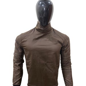 May include: Brown long-sleeved shirt and trousers set with white stitching details. The shirt has a high collar and black cuffs. The trousers have a matching brown colour and white stitching. The outfit is displayed on a mannequin.