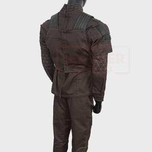 May include: Brown tactical jacket and pants set. The jacket features a high collar, shoulder padding, and geometric stitching on the sleeves. White stitching outlines the seams. The pants have a matching color and style. The outfit is displayed on a mannequin.