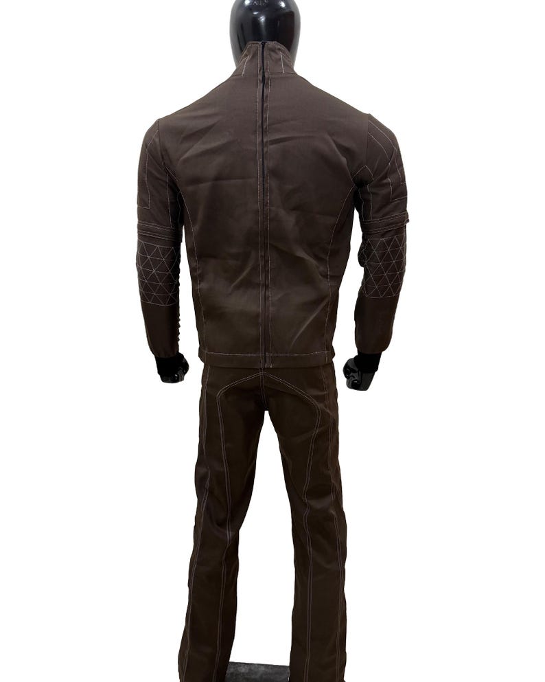 Może przedstawiać: Brązowy zestaw kurtka i spodnie. Kurtka ma wysoki kołnierz i białe przeszycia. Spodnie mają proste nogawki z pasującymi przeszyciami. Rękawy kurtki mają geometryczny wz&oacute;r na ramieniu.