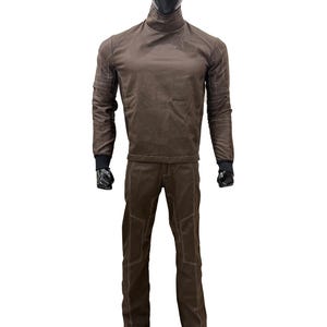 Może przedstawiać: Brązowy dwuczęściowy str&oacute;j na manekinie. G&oacute;ra to koszula z długim rękawem z golfem i białymi przeszyciami. Spodnie są brązowe z białymi przeszyciami i nakolannikami. Manekin ma czarną głowę i dłonie.