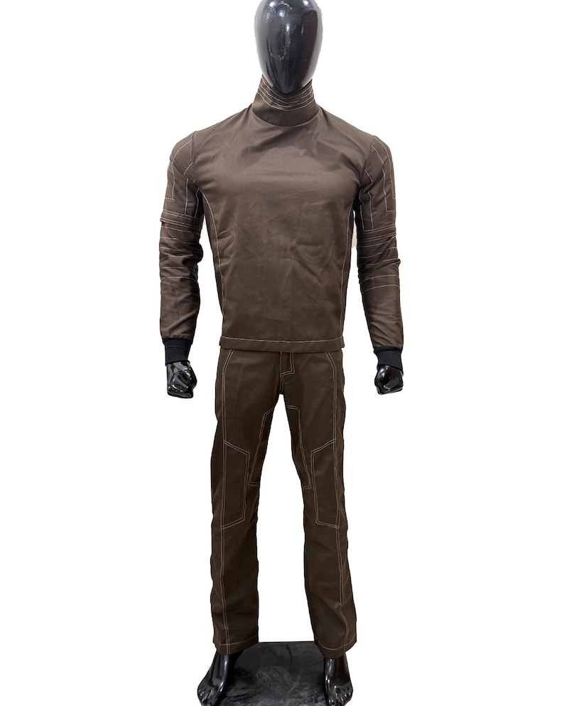 Może przedstawiać: Brązowy dwuczęściowy str&oacute;j na manekinie. G&oacute;ra to koszula z długim rękawem z golfem i białymi przeszyciami. Spodnie są brązowe z białymi przeszyciami i nakolannikami. Manekin ma czarną głowę i dłonie.