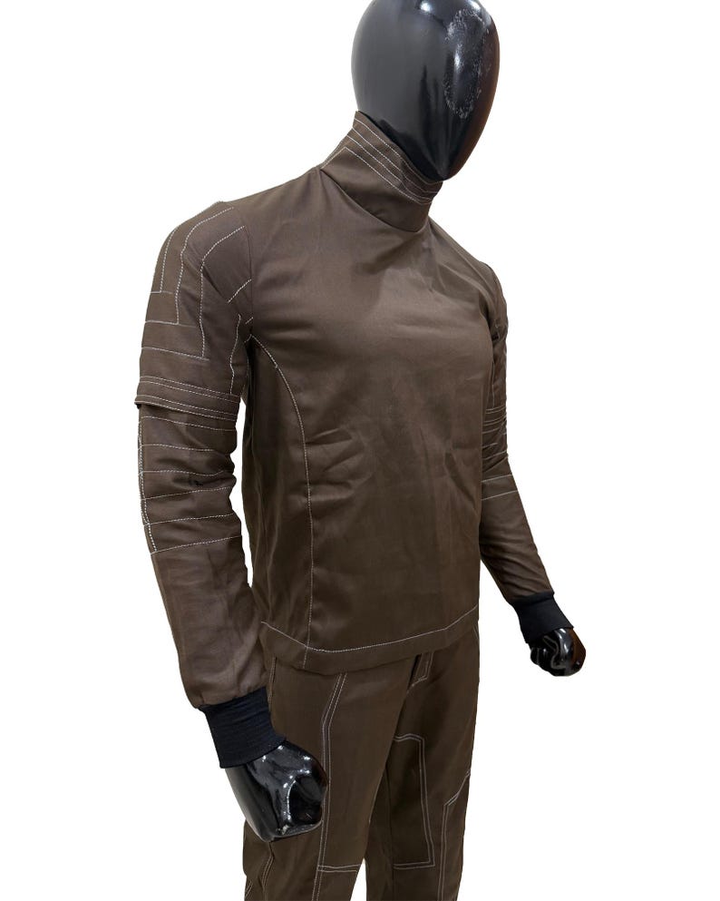 Może przedstawiać: Brązowy kombinezon z białymi detalami szw&oacute;w. Str&oacute;j posiada wysoki kołnierz, długie rękawy i pasujące spodnie. Rękawy mają czarne mankiety. Głowa manekina jest czarna.