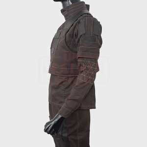 May include: A brown tactical outfit with a high collar and multiple panels. The outfit features white stitching and geometric patterns on the sleeves. The mannequin is wearing a black head covering and gloves.