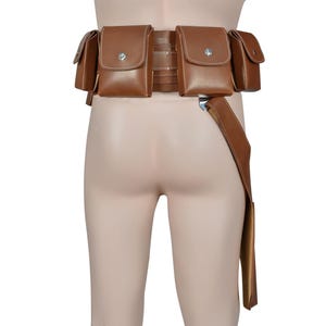 K&ouml;nnte beinhalten: Ein brauner Leder-Werkzeugg&uuml;rtel mit mehreren Taschen und einem langen Riemen. Der G&uuml;rtel hat mehrere quadratische Taschen mit silbernen Knopfverschl&uuml;ssen. Der Riemen h&auml;ngt auf der rechten Seite herunter. Der G&uuml;rtel ist zum Tragen um die Taille konzipiert.
