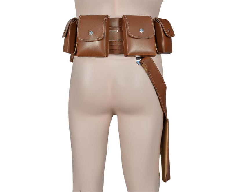 K&ouml;nnte beinhalten: Ein brauner Leder-Werkzeugg&uuml;rtel mit mehreren Taschen und einem langen Riemen. Der G&uuml;rtel hat mehrere quadratische Taschen mit silbernen Knopfverschl&uuml;ssen. Der Riemen h&auml;ngt auf der rechten Seite herunter. Der G&uuml;rtel ist zum Tragen um die Taille konzipiert.