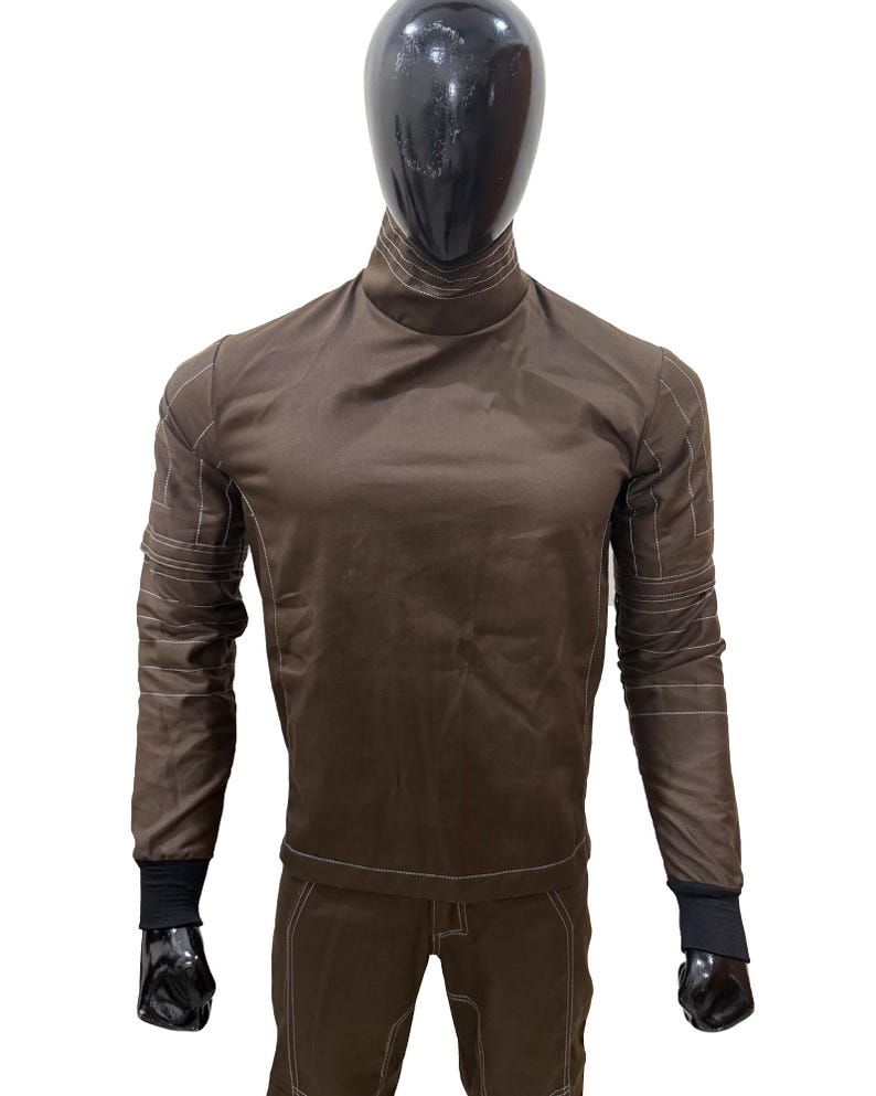 May include: Brown long-sleeved shirt and trousers set with white stitching details. The shirt has a high collar and black cuffs. The trousers have a matching brown colour and white stitching. The outfit is displayed on a mannequin.