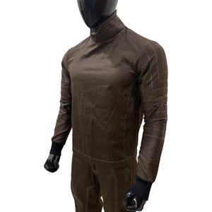 Może przedstawiać: Brązowy kombinezon z wysokim kołnierzem i białymi detalami szw&oacute;w. Top z długim rękawem i spodnie wykonane są z gładkiej tkaniny. Czarne mankiety są widoczne na nadgarstkach i kostkach. Str&oacute;j jest prezentowany na manekinie.