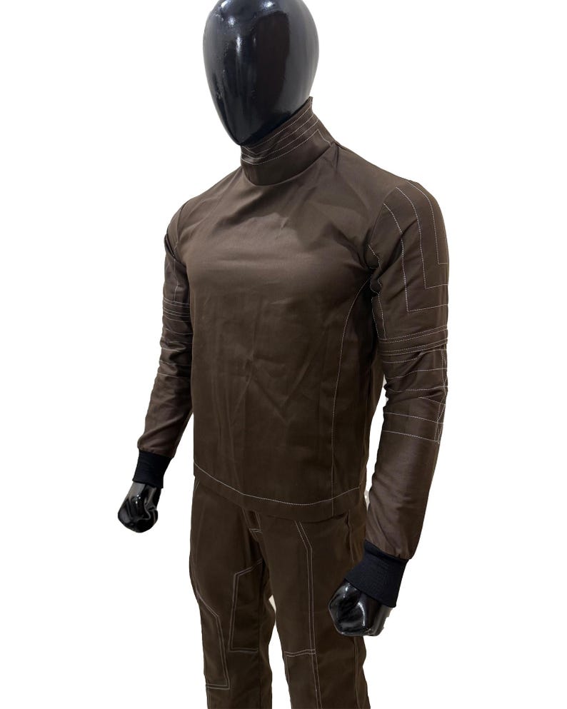 Może przedstawiać: Brązowy kombinezon z wysokim kołnierzem i białymi detalami szw&oacute;w. Top z długim rękawem i spodnie wykonane są z gładkiej tkaniny. Czarne mankiety są widoczne na nadgarstkach i kostkach. Str&oacute;j jest prezentowany na manekinie.