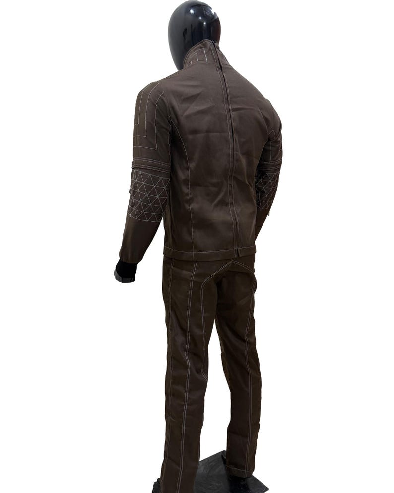 Może przedstawiać: Brązowy dwuczęściowy garnitur prezentowany na manekinie. Kurtka ma geometryczne przeszycia na rękawach i ramionach. Spodnie mają pasujące przeszycia. Garnitur jest wykonany z wytrzymałego materiału.