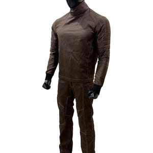 Może przedstawiać: Brązowy dwuczęściowy str&oacute;j na manekinie. G&oacute;ra to koszula z długim rękawem i wysokim kołnierzem z białymi przeszyciami. Spodnie są brązowe z białymi przeszyciami i wzorem paneli. Manekin ma czarną głowę i czarne rękawice.