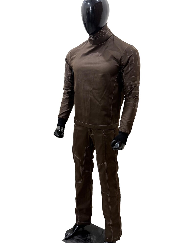 Może przedstawiać: Brązowy dwuczęściowy str&oacute;j na manekinie. G&oacute;ra to koszula z długim rękawem i wysokim kołnierzem z białymi przeszyciami. Spodnie są brązowe z białymi przeszyciami i wzorem paneli. Manekin ma czarną głowę i czarne rękawice.