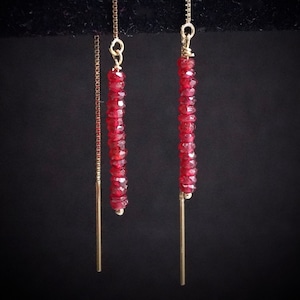 May include: Gold threader earrings with a vertical design. Each earring features a strand of small, faceted red beads. The earrings hang from a delicate gold chain and a straight gold bar.
