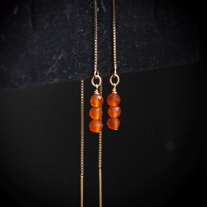 Puede incluir: Pendientes colgantes dorados. Cada pendiente presenta tres cuentas facetadas de color naranja suspendidas de una fina cadena. El fondo oscuro resalta el cálido color de las piedras preciosas.