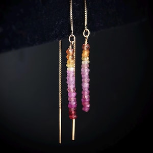 May include: Threader earrings in gold tone, adorned with a cascade of colourful gemstone beads. The earrings showcase a vertical arrangement of small, round beads in pink, orange, yellow, and red, suspended from a fine gold chain.