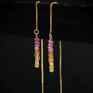 May include: A pair of gold threader earrings. Each earring has a chain with a vertical strand of colourful gemstones in shades of pink, orange, and yellow. The earrings hang against a black background.