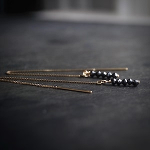 May include: Close-up of delicate gold chain earrings with black beads. The earrings are displayed on a dark surface, highlighting their linear design and the contrast between the gold and black elements. The focus is on the jewellery.