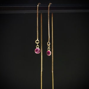 Pode incluir: Um par de brincos de corrente de ouro com pequenas pedras preciosas cor-de-rosa no final de cada corrente.