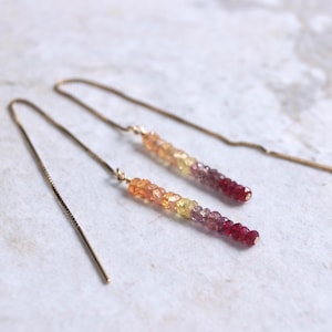 May include: A pair of gold threader earrings. Each earring has a gradient of small, colourful beads, changing from orange to yellow, pink, and then deep red. The earrings are shown on a light-coloured, textured surface.
