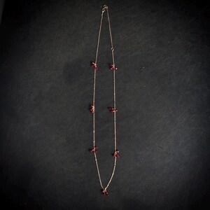 May include: A delicate gold-toned necklace with small, clustered red beads. The necklace features a fine chain and is displayed against a dark background. The beads are evenly spaced along the chain, creating an elegant and minimalist design.