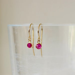 Puede incluir: Un par de pendientes colgantes de oro con pequeñas piedras preciosas rosas, redondas y facetadas, en un engaste sencillo. Los pendientes cuelgan de finos ganchos dorados curvados, mostrando un diseño minimalista con acabado metálico.