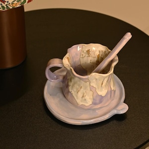May include: A ceramic teacup and saucer set in shades of lavender and cream. The cup has a unique, irregular shape with a handle, and a matching spoon rests inside. The set is placed on a dark surface, showcasing the handcrafted details.