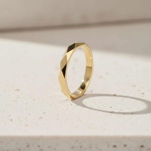 Könnte beinhalten: Ein goldener Ring mit einem geometrischen, facettierten Design. Der Ring hat ein Band mit mehreren flachen Oberflächen, wodurch ein strukturiertes Aussehen entsteht. Der Ring ist vor einem neutralen Hintergrund platziert, der seine polierte, reflektierende Oberfläche hervorhebt.