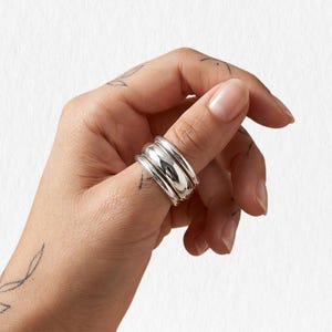 Könnte beinhalten: Ein breiter, silberner Ring mit einem abgerundeten, geschichteten Design. Der Ring wird an einem Finger getragen und zeigt seine polierte, reflektierende Oberfläche. Das Schmuckstück hat eine moderne, minimalistische Ästhetik.